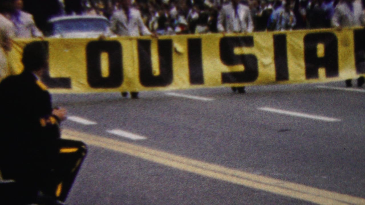 1961 California 8mm frame showing a large yellow LOUISIANA banner crossing a street, with a dense crowd behind it and a suited spectator at the left edge.