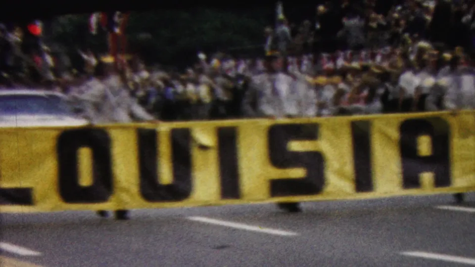 1961 California home-movie frame showing the lower portion of a yellow LOUISIANA banner across the street, with spectators and a light-colored car visible in the background.
