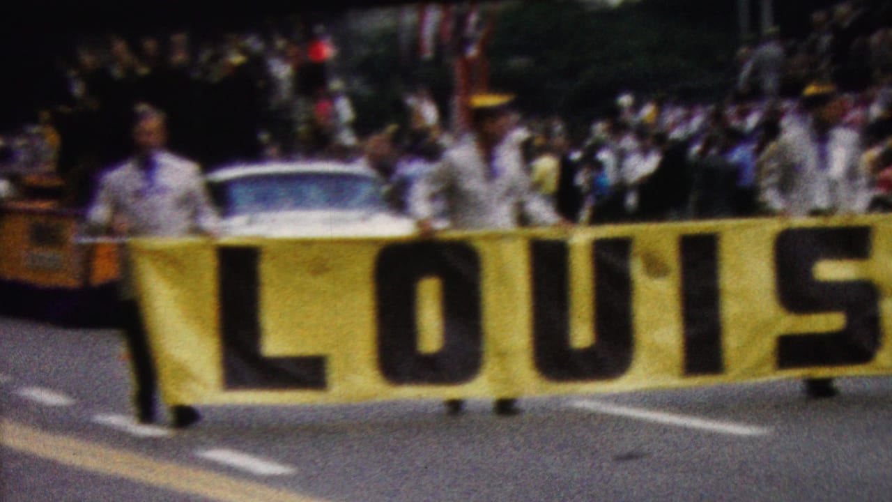 1961 California parade frame showing uniformed participants carrying a large yellow LOUISIANA banner, with spectators and a white car visible behind them.