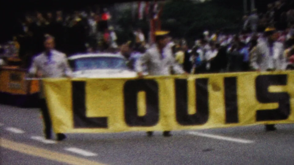 1961 California parade frame showing uniformed participants carrying a large yellow LOUISIANA banner, with spectators and a white car visible behind them.
