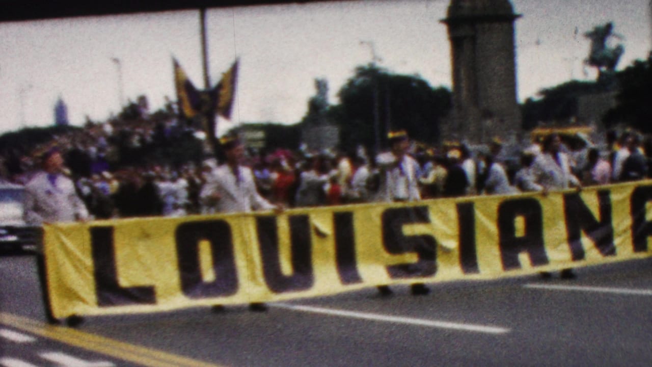 Louisiana parade banner stretches across the road as marchers and spectators fill the background.