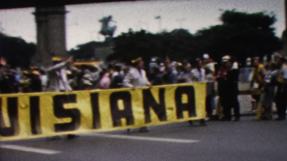 1961 California parade frame showing the lower portion of the LOUISIANA banner crossing the foreground, with spectators, uniformed marchers, and a tall background structure visible.