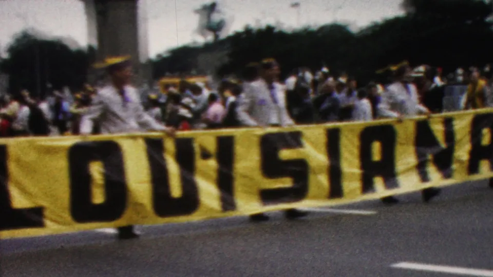 1961 California parade frame with the yellow LOUISIANA banner close to the camera, uniformed participants behind it, and spectators gathered along the street.