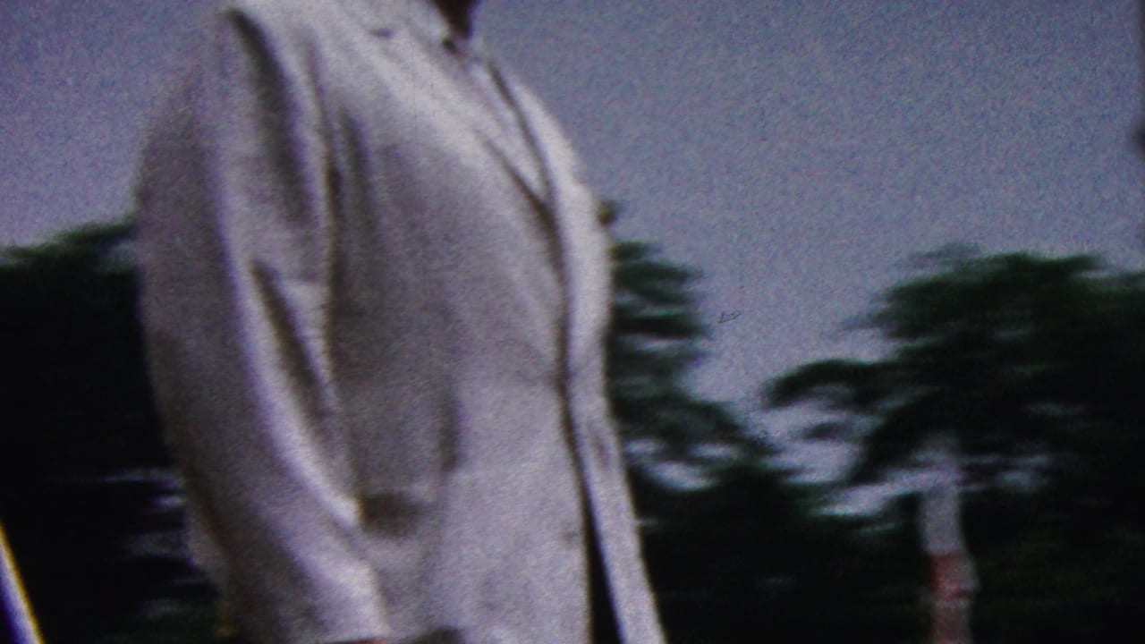 A man in a white suit is partially framed while walking along a parade route in 1961 California, with trees and motion blur behind him.