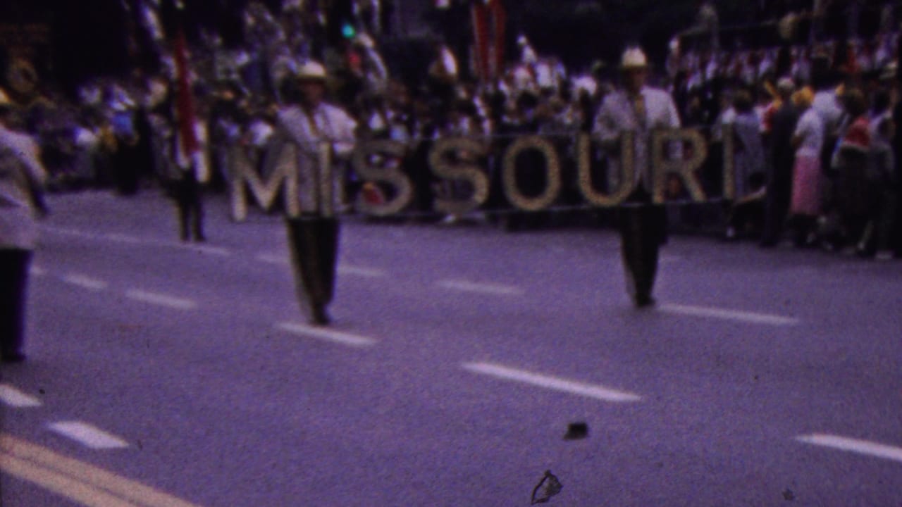 In supported 1961 California parade footage, two men carry a large MISSOURI sign down a broad street while spectators line the curb in the background.
