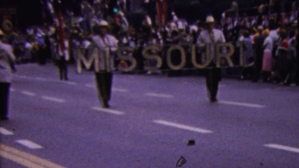 In supported 1961 California parade footage, two men carry a large MISSOURI sign down a broad street while spectators line the curb in the background.