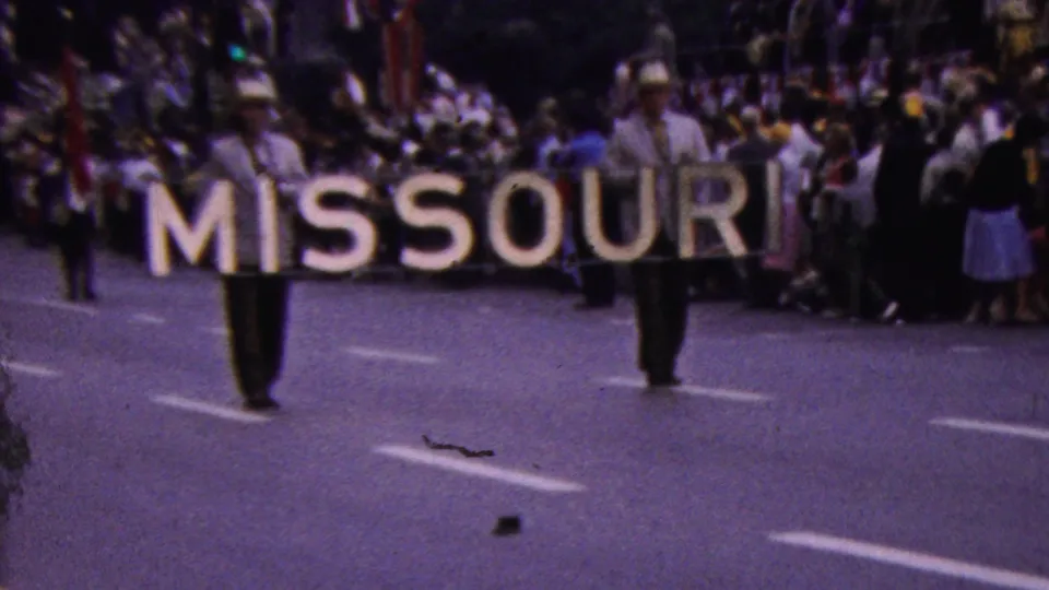 Two parade participants in light shirts and hats carry the large MISSOURI lettering past a crowded sidewalk, with additional parade members and spectators behind them.
