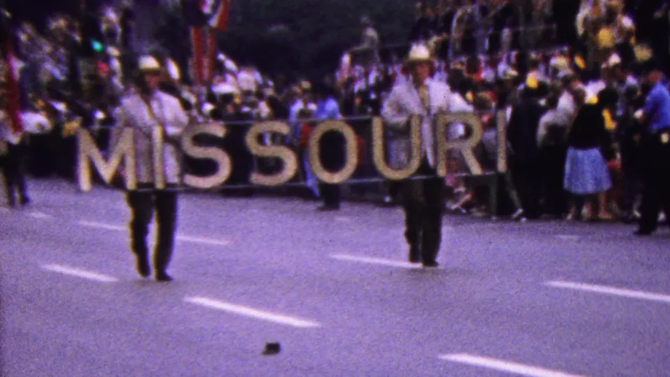 Marching-band members in light uniforms and cowboy-style hats appear near the MISSOURI sign as the procession advances past spectators on the street.