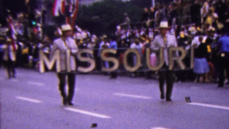 A soft-focus 8mm frame shows marching musicians in light uniforms with hats beside the large MISSOURI sign, with a dense spectator crowd and flags behind them.