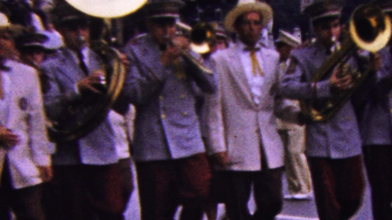 Uniformed marching band members in 1961 California move toward the camera with brass instruments and red trousers visible in a crowded parade scene.
