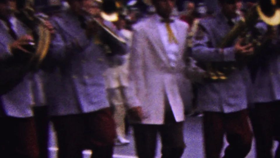 A close, slightly blurred 8mm view shows uniformed male musicians with tubas and horns marching past the camera in California in 1961.