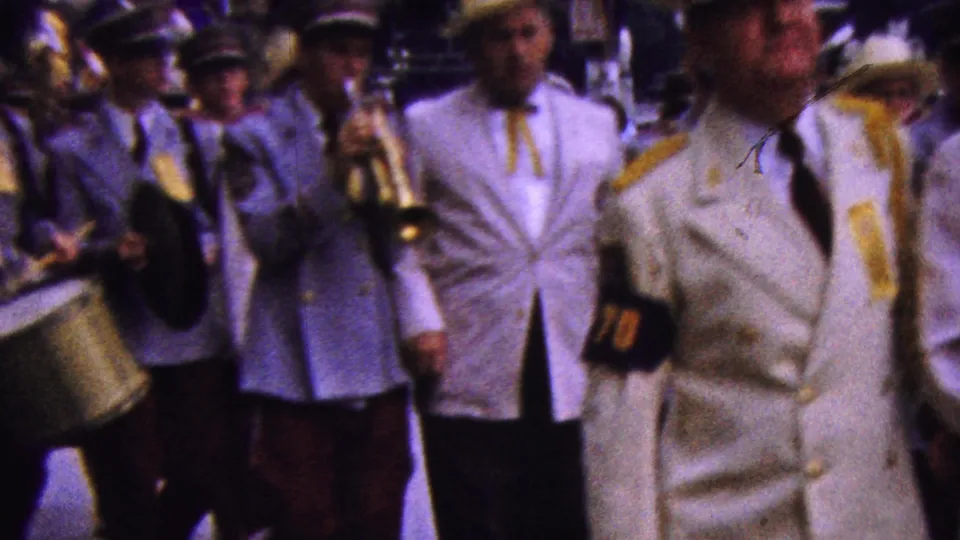 A man in a white parade suit and straw hat walks ahead of uniformed musicians with brass instruments and a drum in 1961 California.