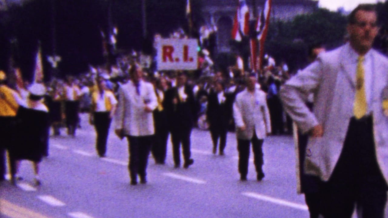 1961 California parade footage showing white-jacketed marchers with yellow ties approaching the camera as an R.I. placard and spectators appear behind them.
