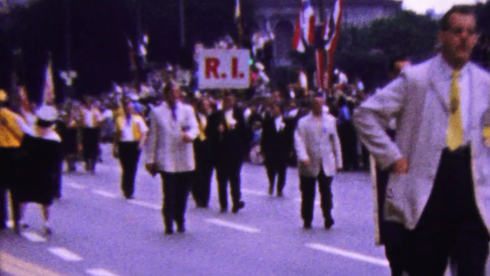 1961 California parade footage showing white-jacketed marchers with yellow ties approaching the camera as an R.I. placard and spectators appear behind them.