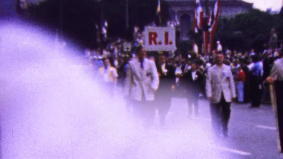 A man in a white jacket and yellow tie stands near the camera during the 1961 California parade, with more marchers, spectators, and an R.I. sign visible in the background.