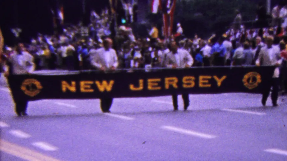 1961 California parade frame with participants carrying a black NEW JERSEY banner in yellow lettering as spectators and additional marchers fill the background.