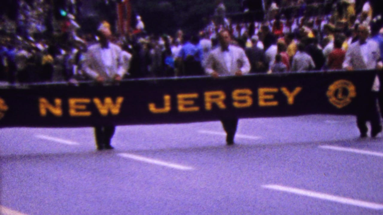 1961 8mm home-movie frame showing men in white shirts carrying a black NEW JERSEY banner with yellow lettering during a crowded street parade in California.