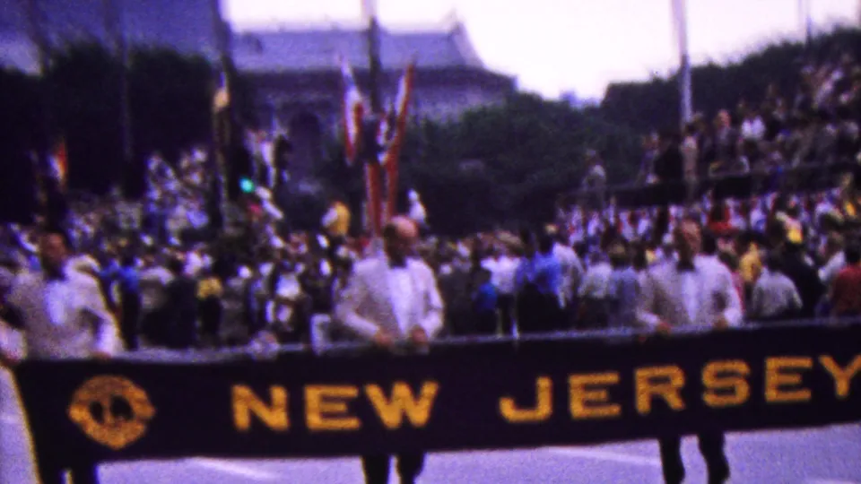 Street-level 8mm frame of a parade group carrying a NEW JERSEY banner, with spectators lining the route and tall urban structures in the distance.