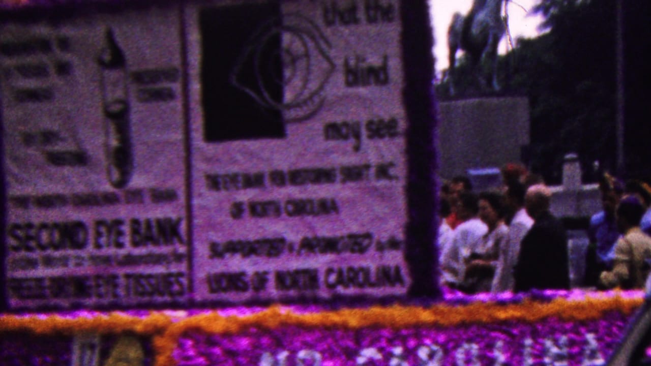 A 1961 California parade float with purple and gold trim passes the camera, showing large eye bank signage and a crowd of onlookers behind it.