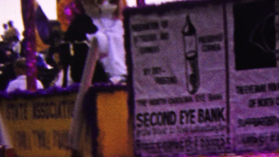 A woman in a white dress stands on the passing 1961 California parade float beside a black dog, with eye bank signage and spectators visible around her.