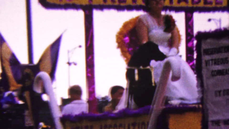 The decorated float fills the frame with swan-like figures, a woman in white, a black dog, purple trim, and spectators partly visible behind the float.