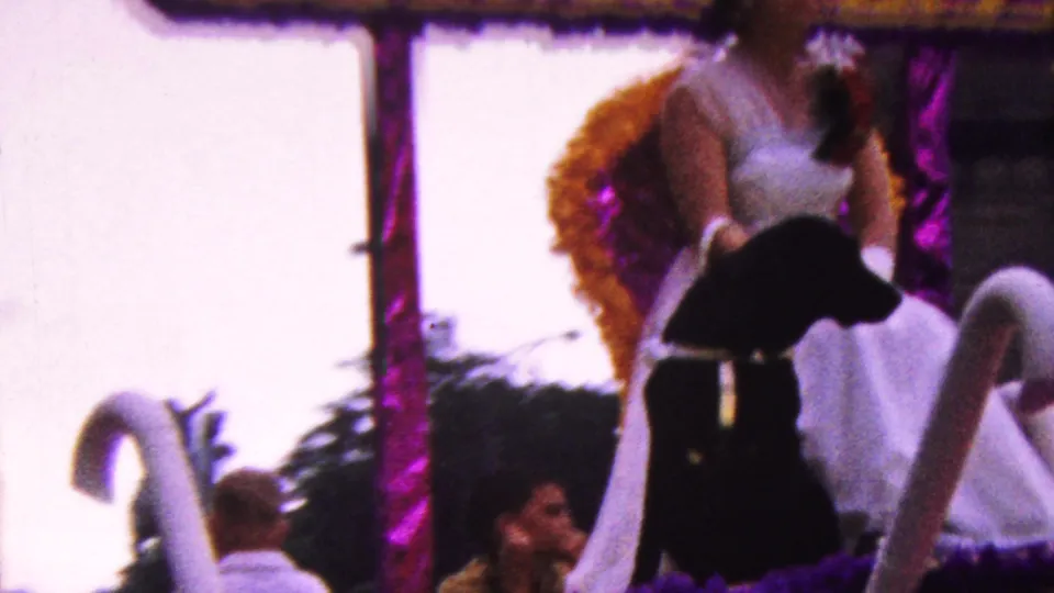 A later frame of the 1961 California parade float shows the woman and black dog near swan-like decorations, with the handheld camera catching spectators behind them.