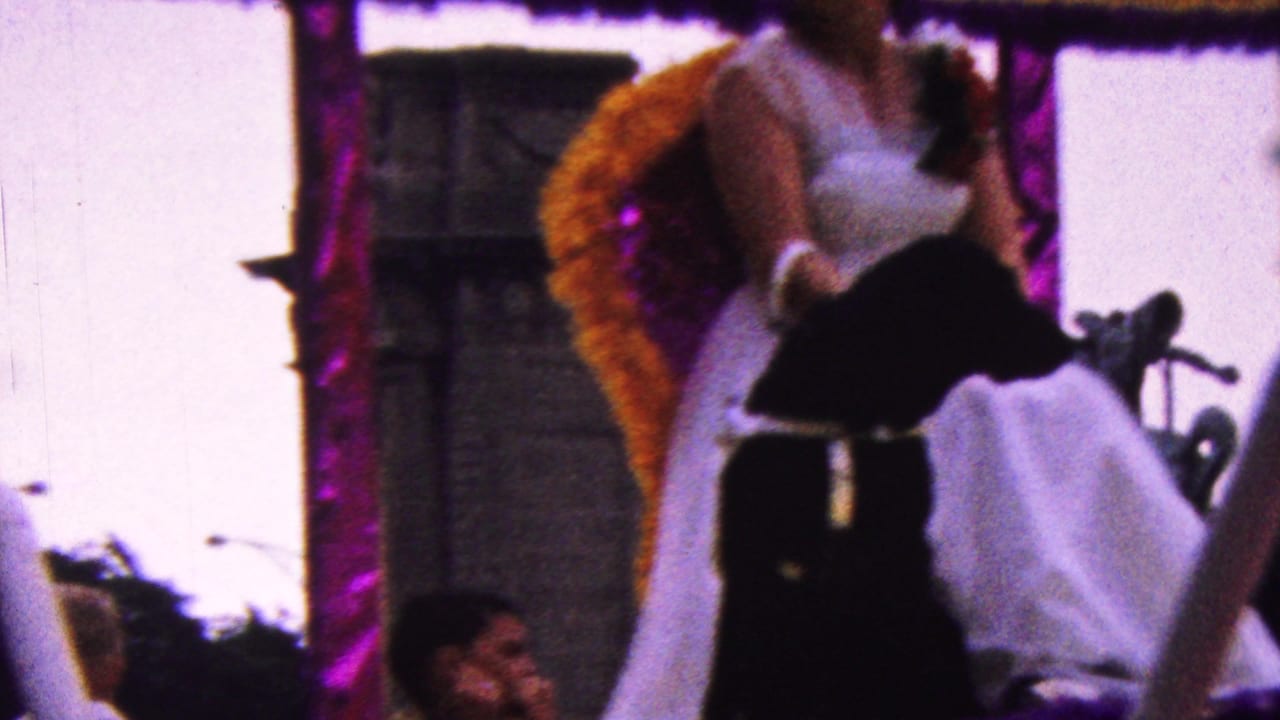 In 1961 California home-movie footage, a woman in a white dress and sunglasses sits on a float with purple and gold decoration as a parade moves along a city street.