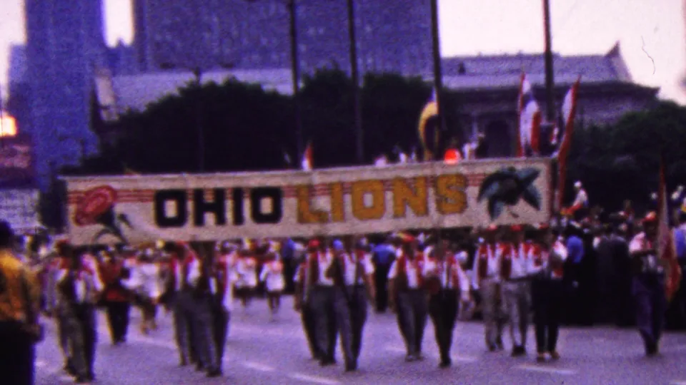 Uniformed parade participants walk toward the camera beneath a large OHIO LIONS banner, with flags and a city building visible behind them.