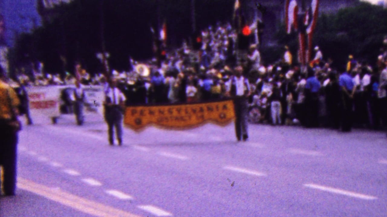 In the supplied 1961 California clip, parade participants carry a yellow Pennsylvania District 14 banner while spectators and a brass band gather farther up the street.