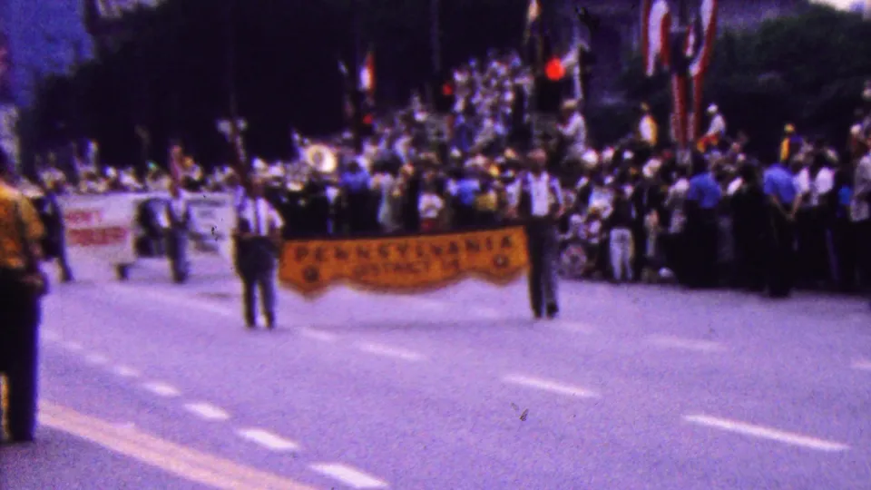 In the supplied 1961 California clip, parade participants carry a yellow Pennsylvania District 14 banner while spectators and a brass band gather farther up the street.
