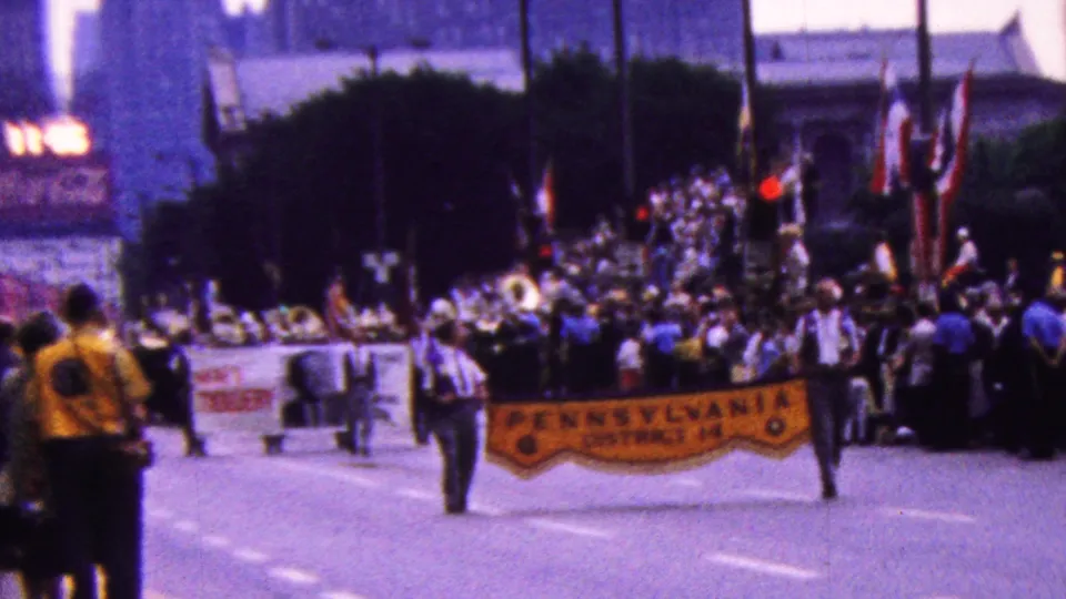 A wider 8mm view shows the parade group on a California street in 1961, with a yellow banner in front, another banner at left, flags raised behind, and spectators clustered along the route.