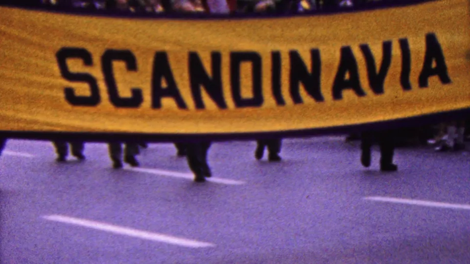 A close 1961 California parade frame shows the yellow banner cropped tightly with the word Scandinavia visible, held above marchers' legs on the street.
