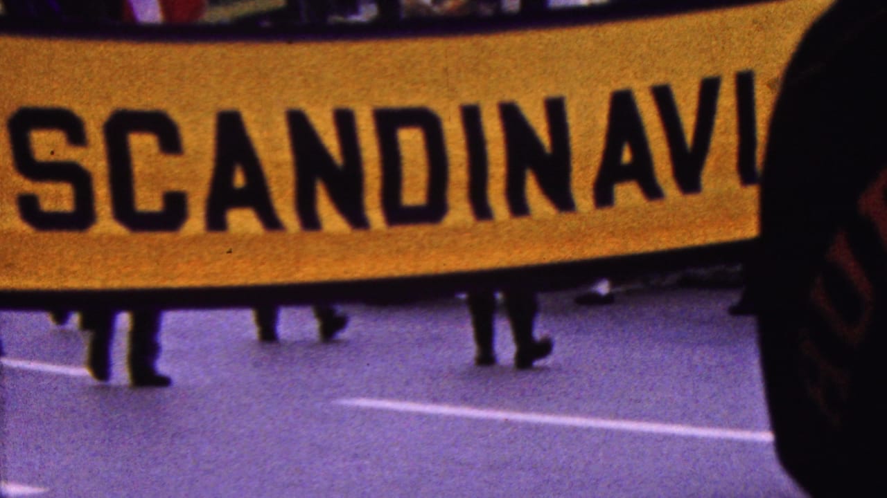 1961 California home-movie frame showing a large yellow SCANDINAVIA banner crossing a paved street, with marchers’ legs and street markings visible below.