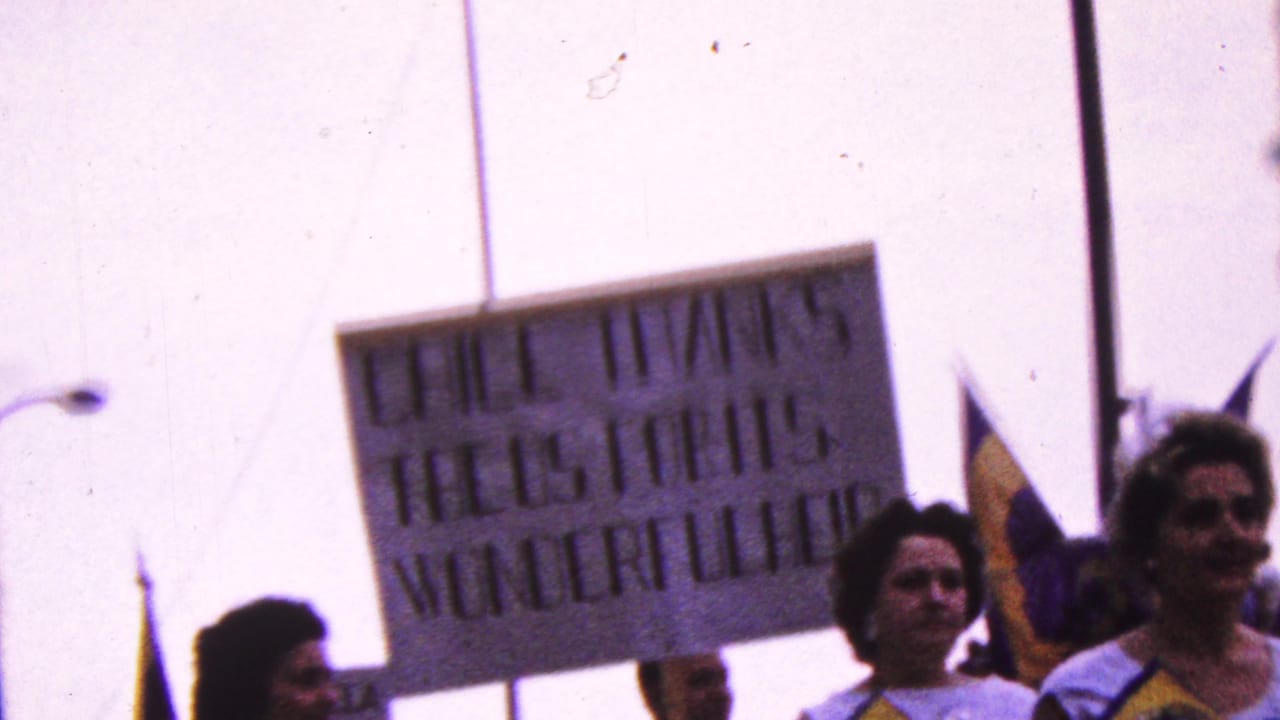 1961 California parade frame with women and other marchers carrying handmade signs and flags, including a partially legible sign and a yellow-and-blue flag at right.