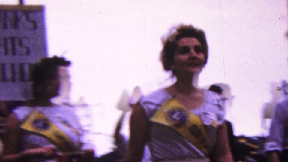 1961 California home-movie frame showing a woman in a white dress and yellow sash walking in a street parade, with another sash-wearing marcher blurred at left.