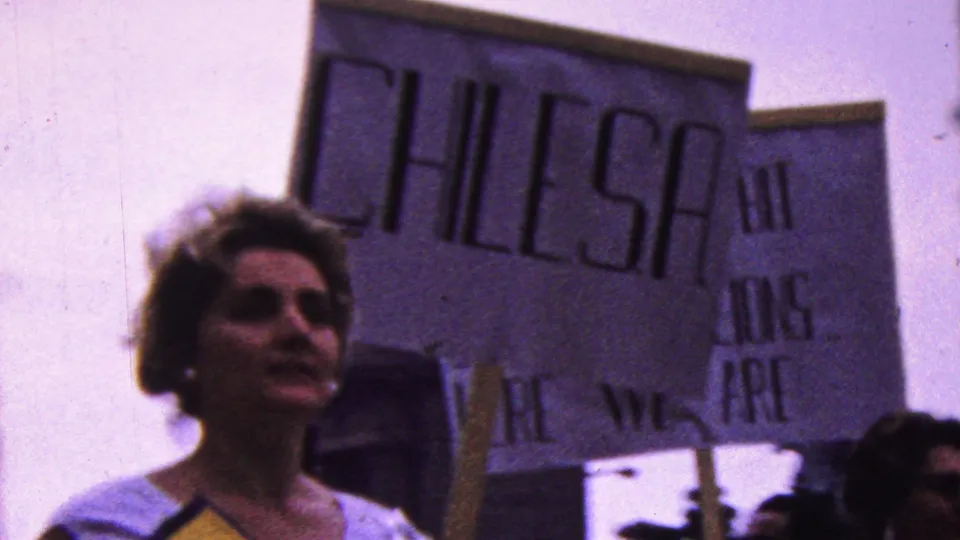 1961 California parade frame showing a woman walking near a large handmade sign with partially legible wording, while other marchers and flags appear at the edges.