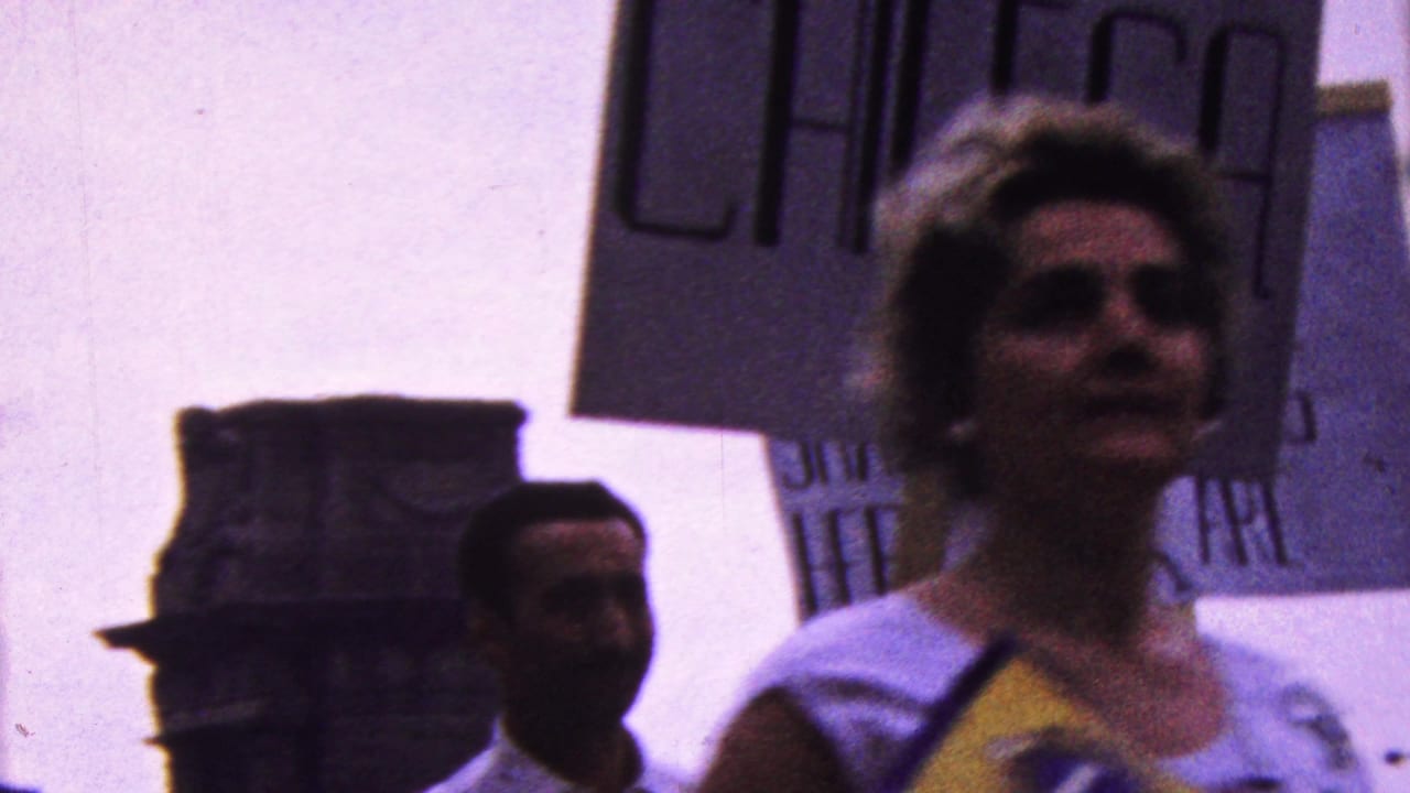 In metadata-supported 1961 California footage, adult marchers pass close to the handheld camera while carrying handmade protest signs, with faces and a building edge visible in the background.