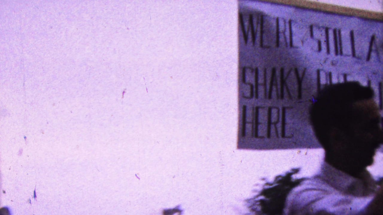 A blonde woman walks in front of protest signs, with another sign partly readable behind her.