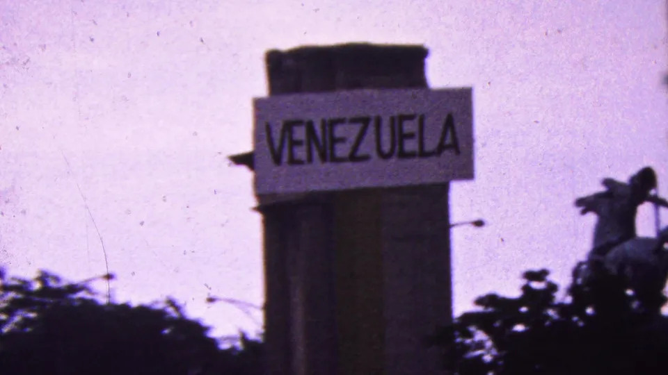 A handmade sign reading VENEZUELA is visible on a vertical structure during the metadata-supported California protest footage, with trees, sky, and a streetlight partly visible nearby.