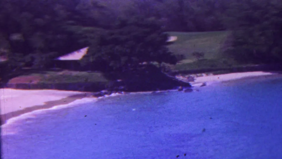 A later 8mm frame continues the Hawaiian coastal overlook, with blue water in the foreground and a pale beach, rocks, trees, and grass along the shore.