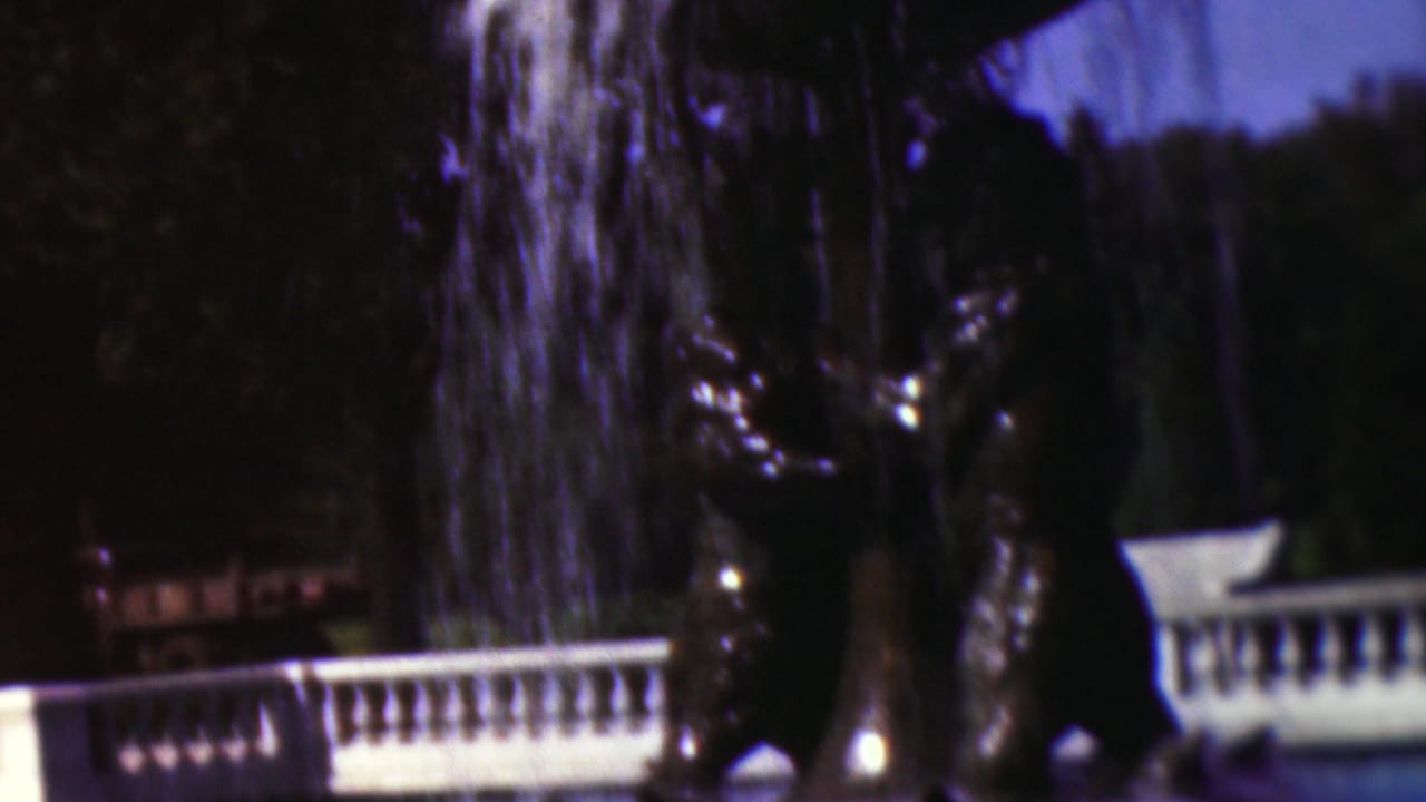 1953 Cleveland 8mm frame showing a dark ornamental fountain statue, falling water, a white balustrade, and shaded trees in an outdoor landscaped setting.