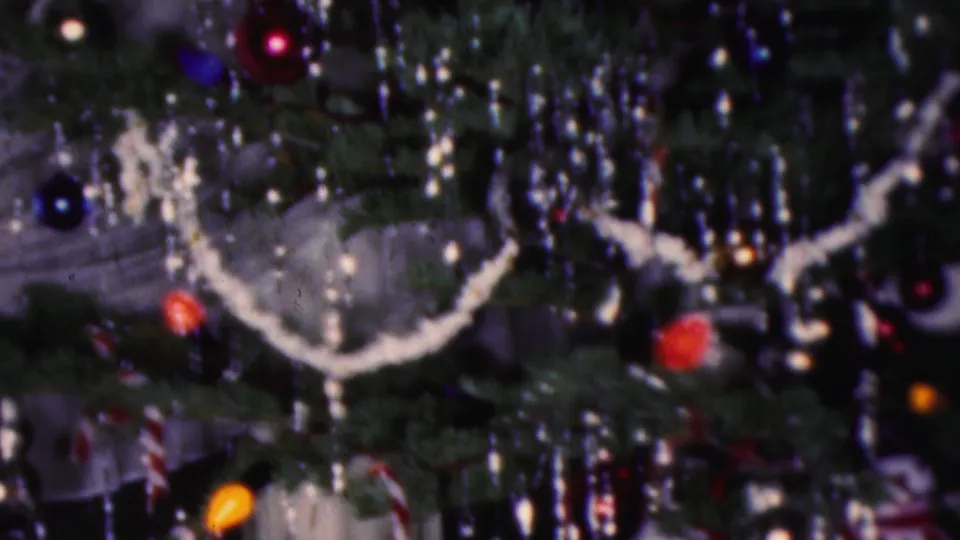 1953 Cleveland home-movie frame showing a dense Christmas tree close-up with reflective tinsel, white garland, red and blue baubles, yellow light glow, and candy-cane striped ornaments.