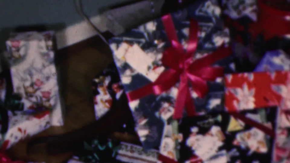 1953 Cleveland home-movie frame showing wrapped Christmas presents clustered around a large red bow, with a wooden furniture leg and patterned paper visible.