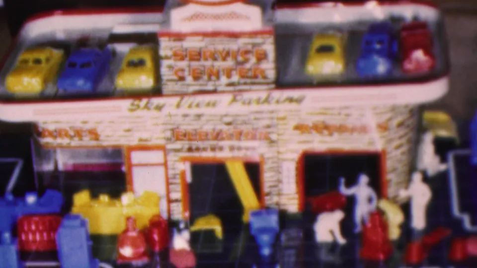 1950s home-movie frame of the toy service center playset with a row of roof cars, service-station signage, plastic figures, and accessories arranged on the dark base.