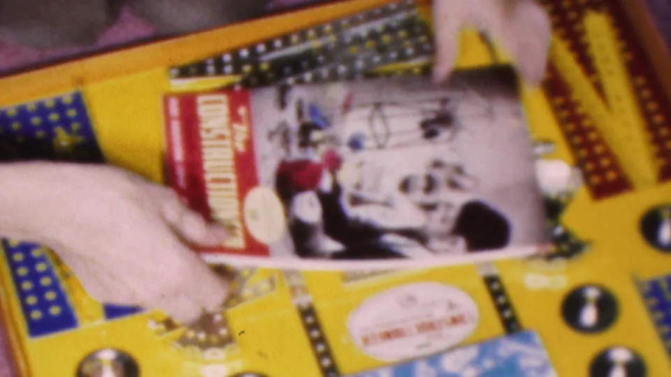 Pale hands handle cards or game pieces on a yellow board-game surface with printed graphics, tokens, and possible branded material in the 1953 Cleveland home-movie clip.