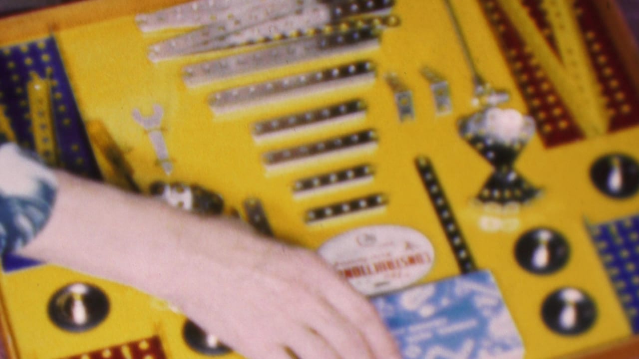 Close 1953 Cleveland home-movie frame showing a hand reaching across a yellow construction set board with small tools, metal hardware, and printed instruction materials.