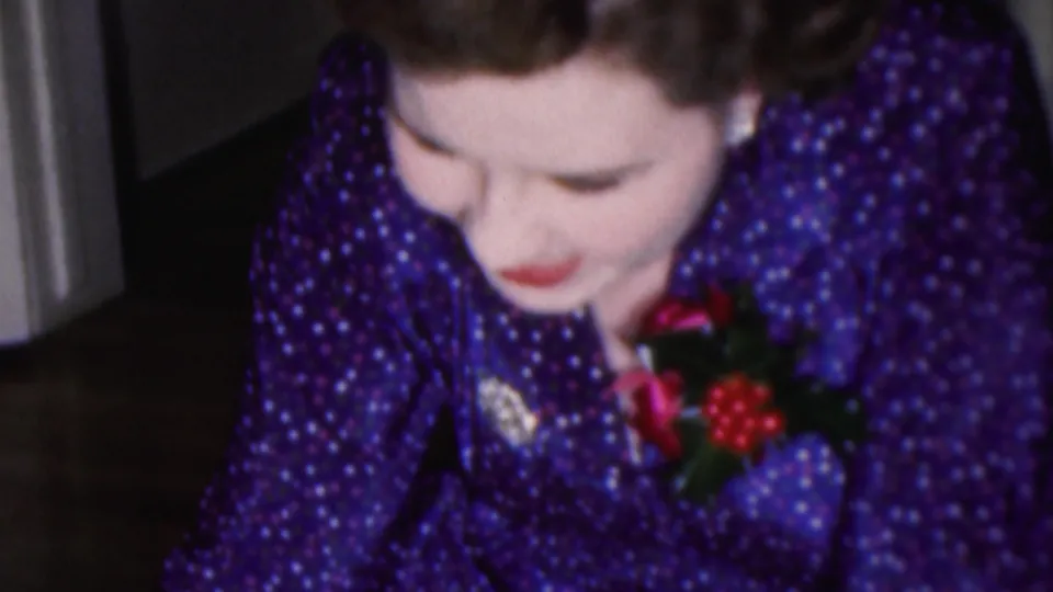 1953 Cleveland home-movie frame of a woman in a purple polka-dot dress with red lipstick and a festive corsage looking down in concentration indoors.