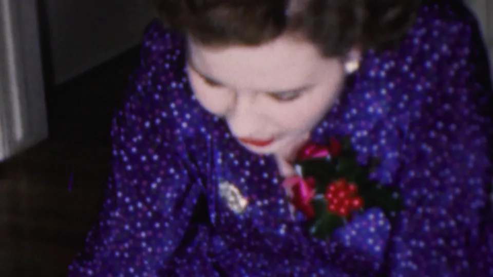 1953 Cleveland home-movie close-up of a woman in a purple polka-dot dress and corsage looking downward as she continues an off-frame assembly activity indoors.