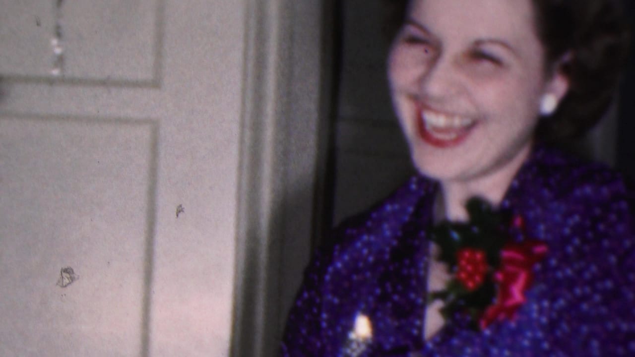 In a 1953 Cleveland home-movie frame, a smiling brunette woman in purple patterned festive clothing stands near a white paneled door with holiday trim behind her.