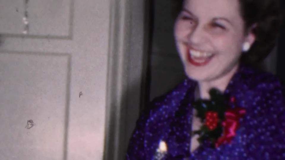 In a 1953 Cleveland home-movie frame, a smiling brunette woman in purple patterned festive clothing stands near a white paneled door with holiday trim behind her.